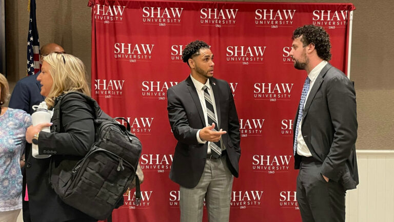 Two men speaking together in front of red Shaw University photo backdrop.